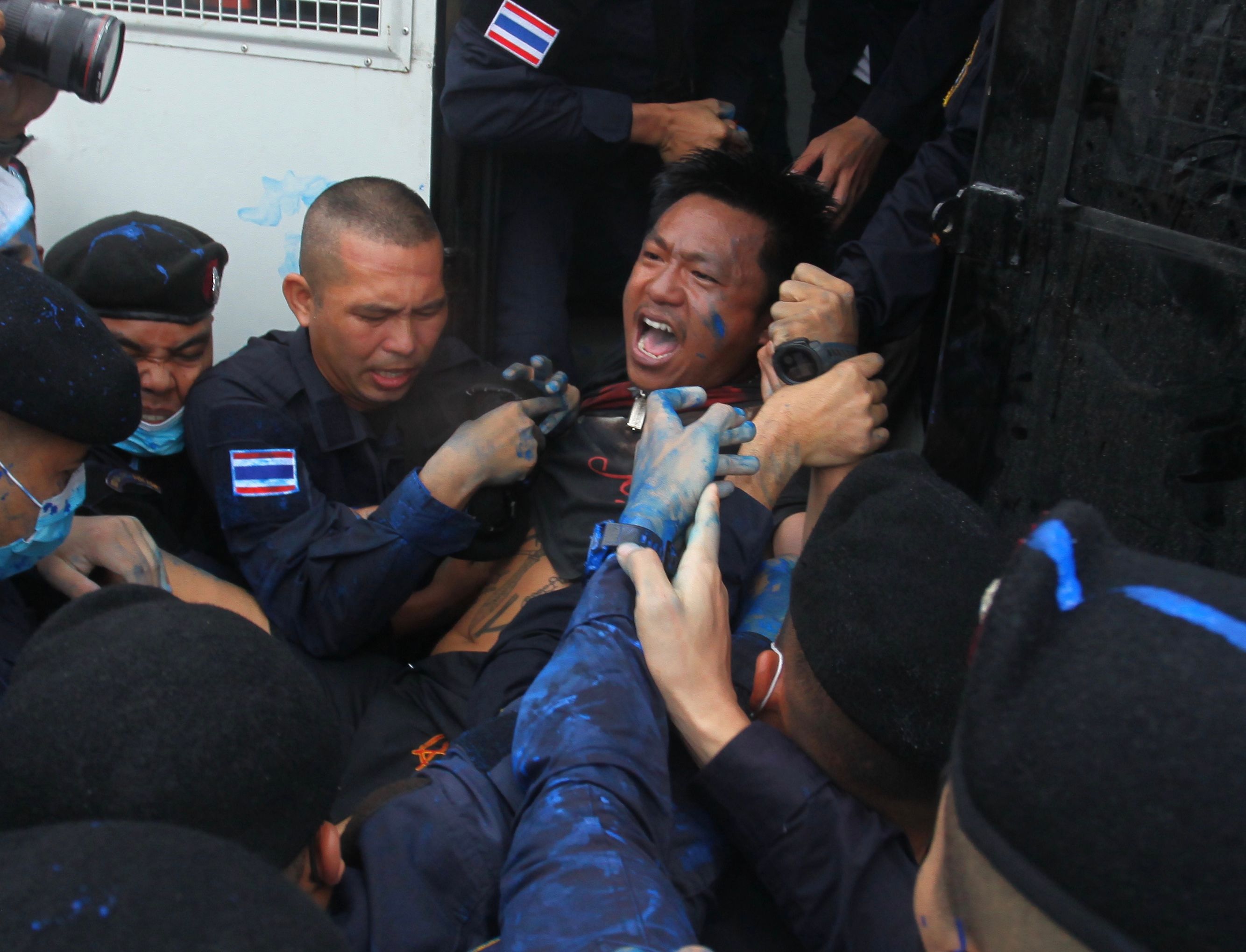 Police arresting a man and bundling him into a police van. They are all stained with blue paint. The man being arrested looks distressed and angry. Some of the police men look aggressive.