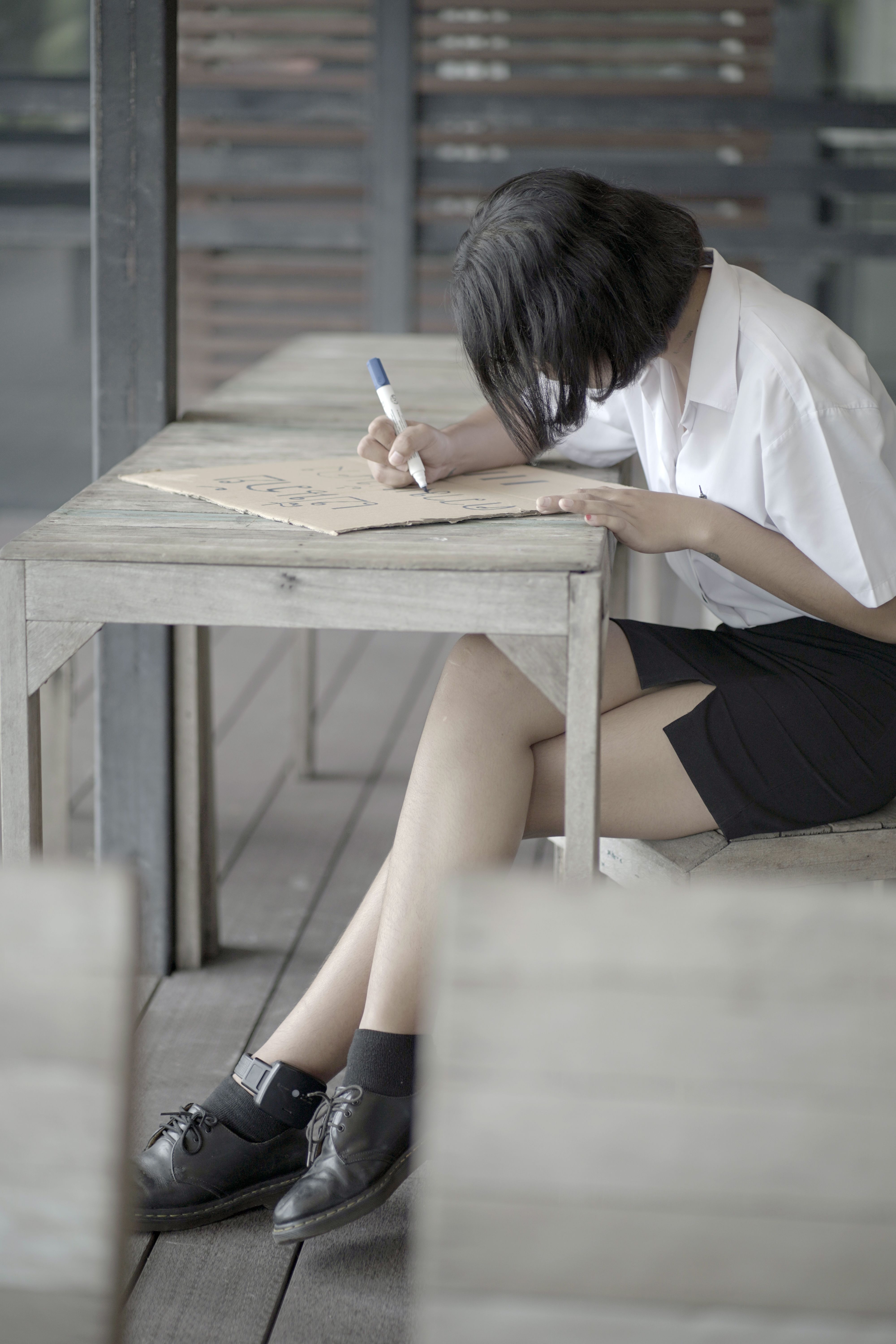 A young Thai woman in a white shirt and black skirt, sitting at a desk and writing with a felt-tip pen on a piece of cardboard. She is making a protest placard.