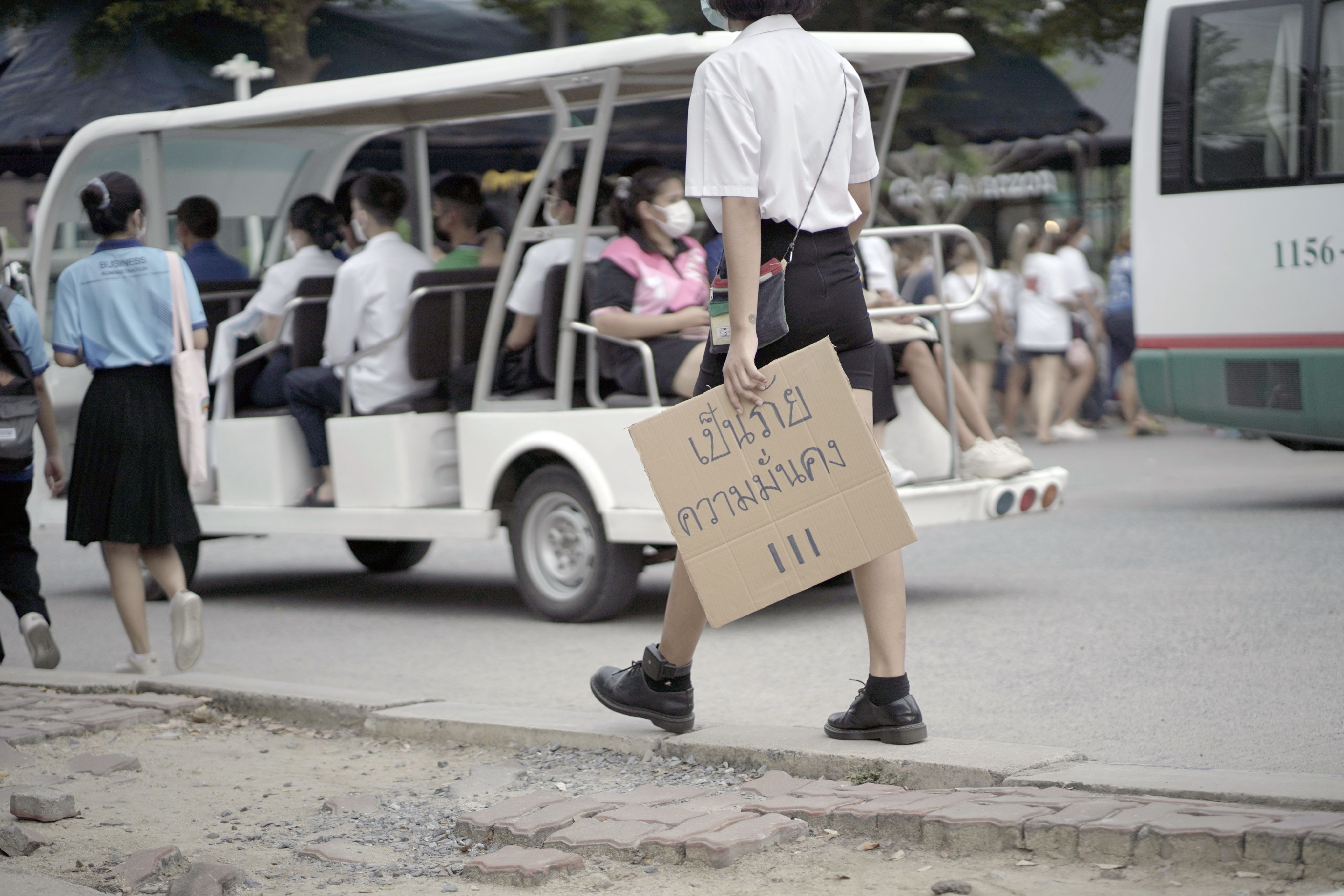The same young Thai woman walking down a busy city street holding the cardboard placard that she's just made. She wears a white shirt, black skirt, and black shoes. The sign says, in Thai, 'Threat to National Security'. She wears a police tagging device around her ankle.