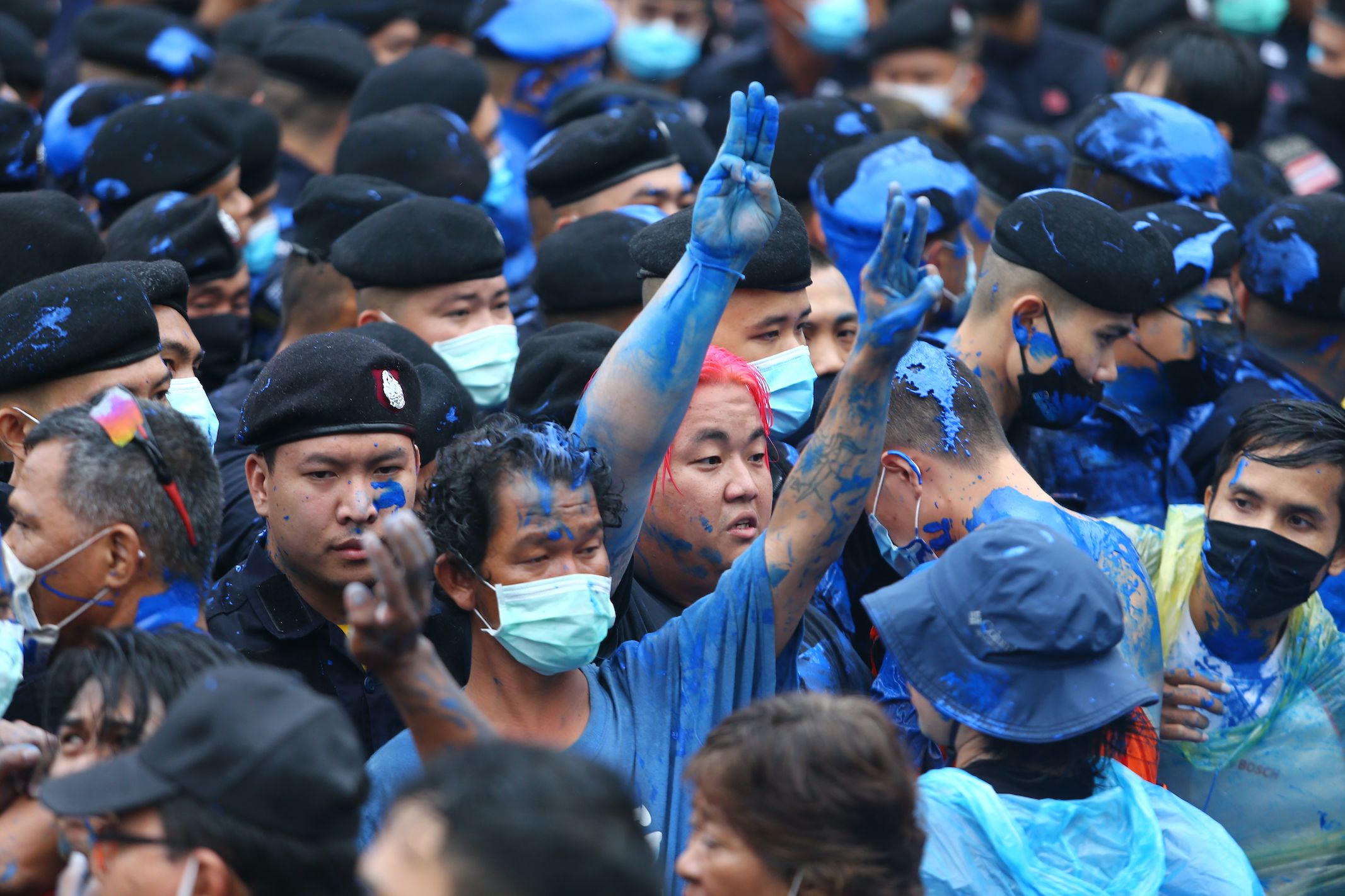 A group of protesters and policemen, some covered in blue paint. Two protesters are giving the three-fingered pro-democracy salute. One is wearing a blue face mask. Another has bright pink hair.