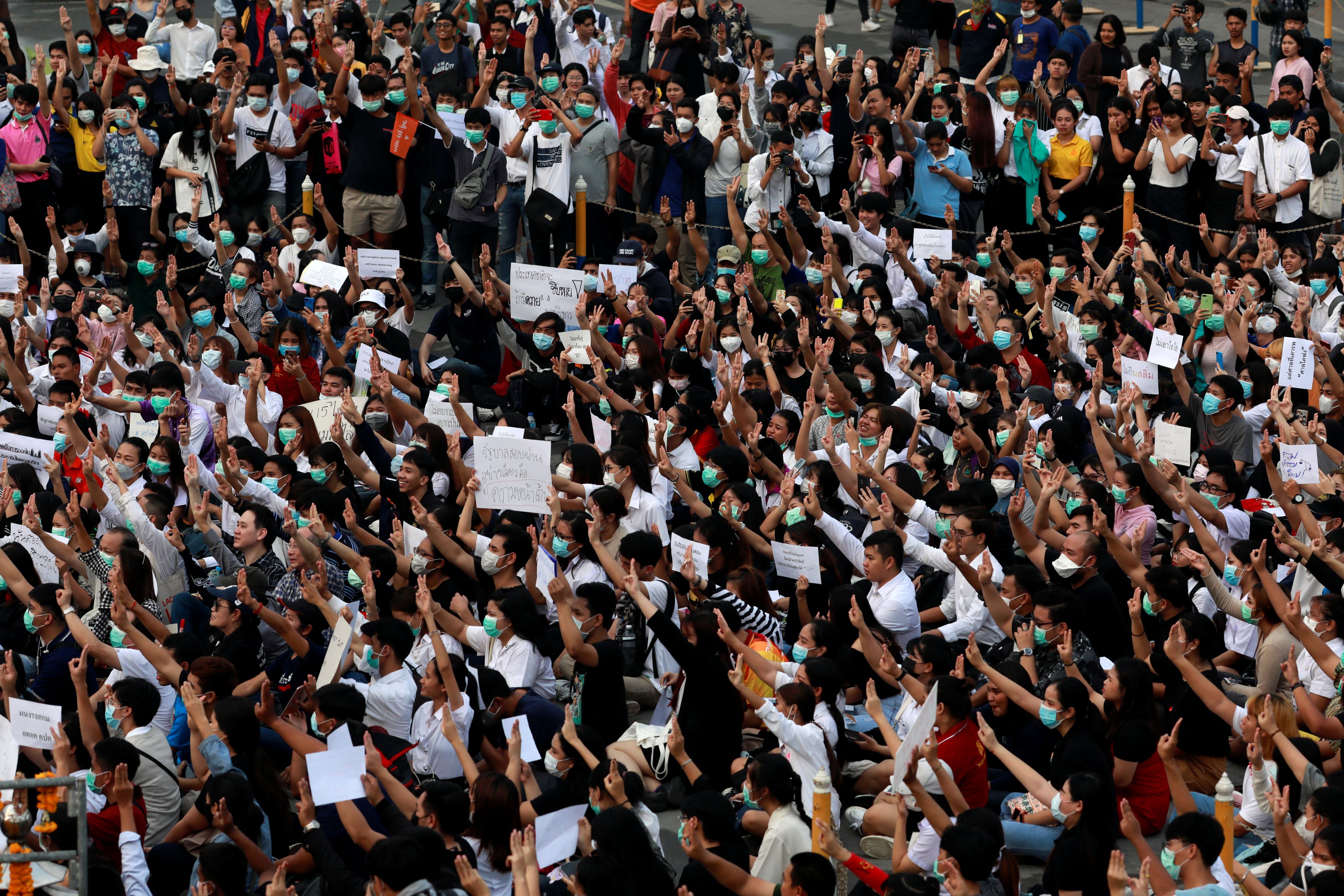 A large group of students at a protest, many making the three-fingered salute and wearing face masks, some holding placards.