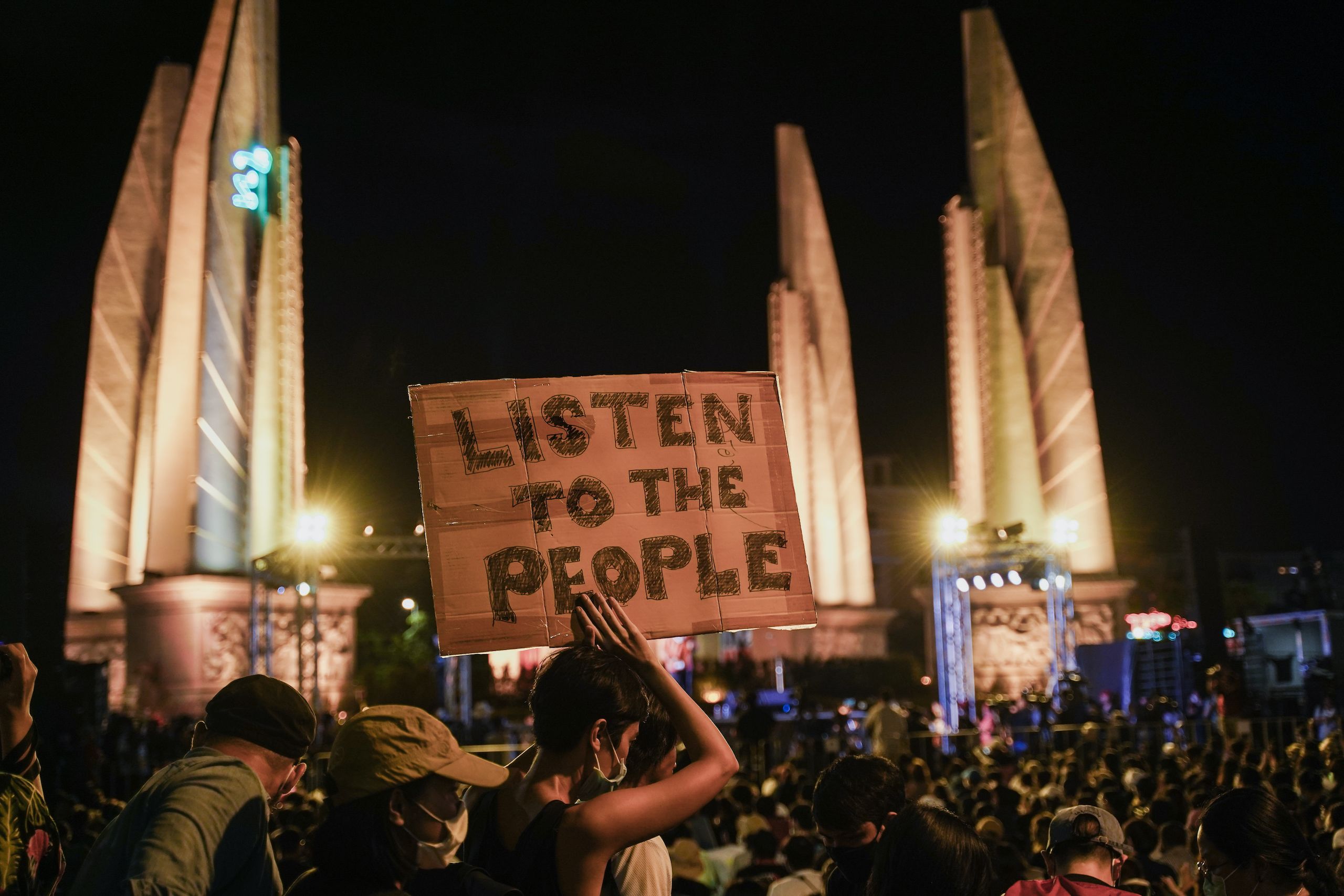 A woman holds a handmade protest sign that reads LISTEN TO THE PEOPLE. In the background is the Democracy Monument of Bangkok. There is a crowd of people around her. It's night time.