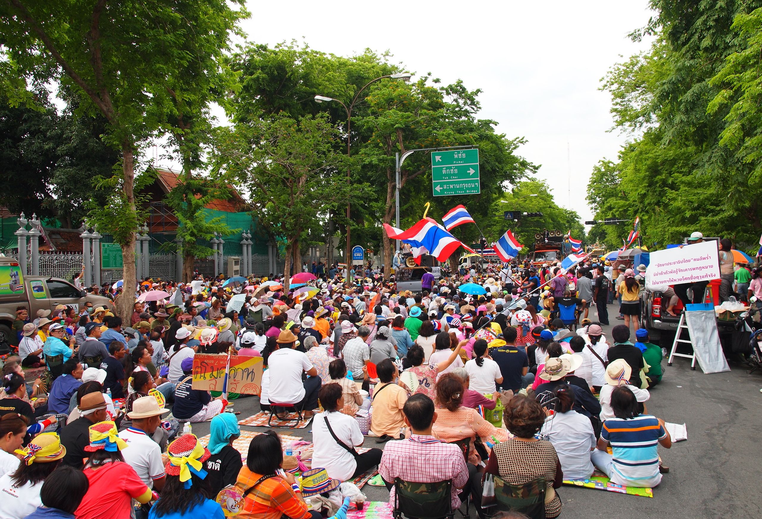 A group of protesters sitting in a city street, facing away from the camera. Some are waving Thailand flags and some carry protest signs.