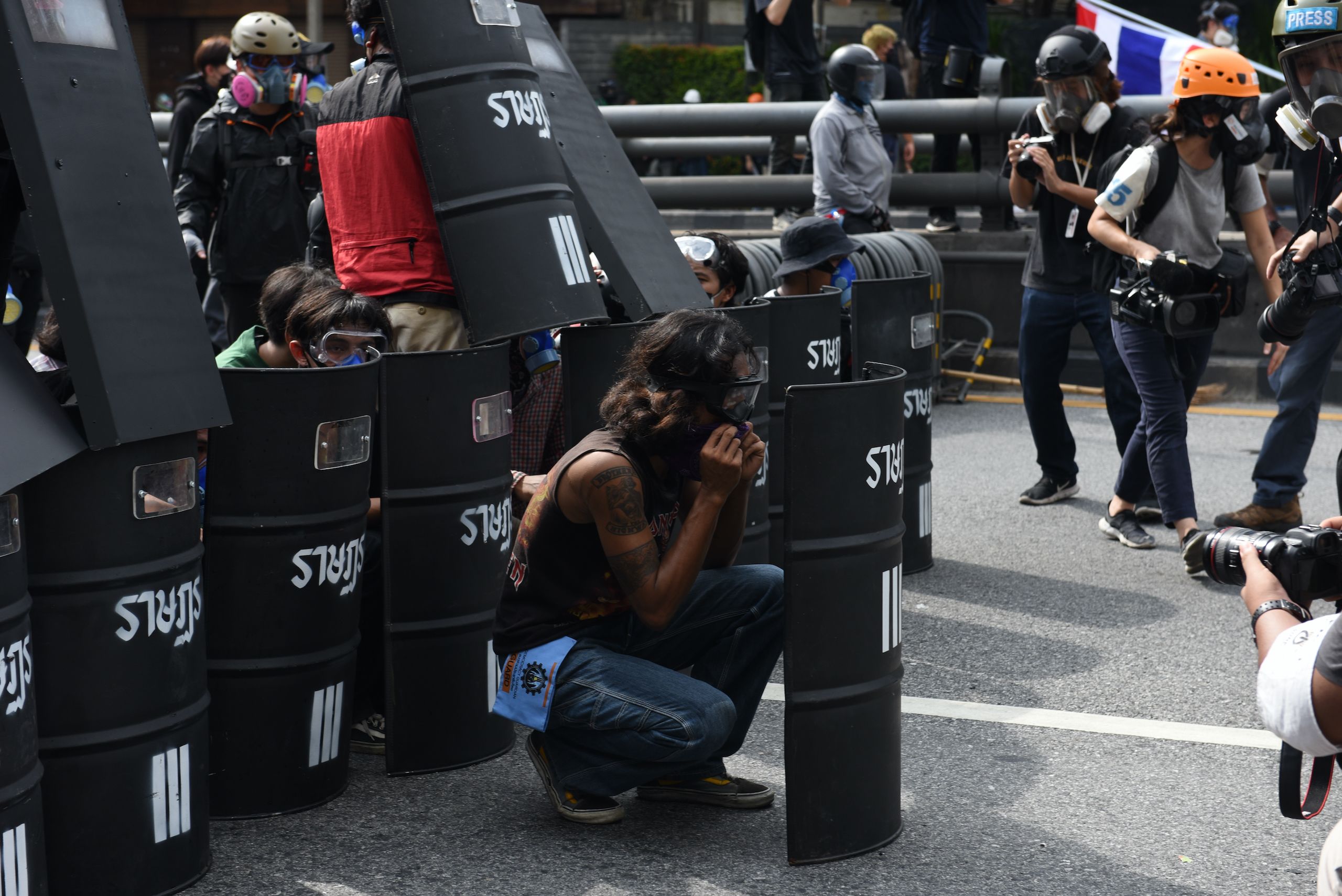 Protesters in the street crouch down behind large black shields. Some wear safety goggles and masks. People are taking photos of them.