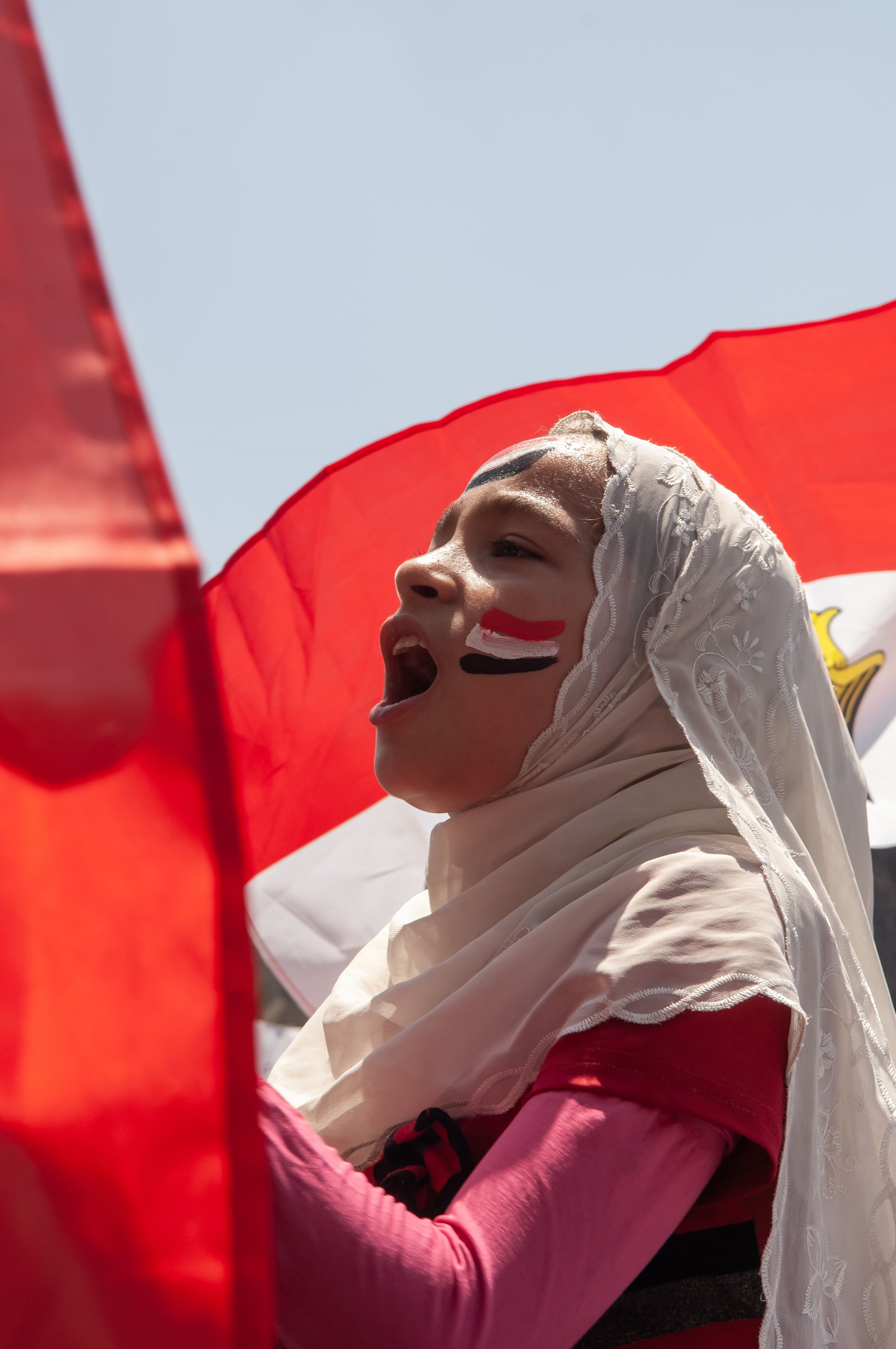 A young Egyptian girl wearing a head scarf, with the colours of the Egyptian flag painted on her cheek, shouting in front of the Egyptian flag. 