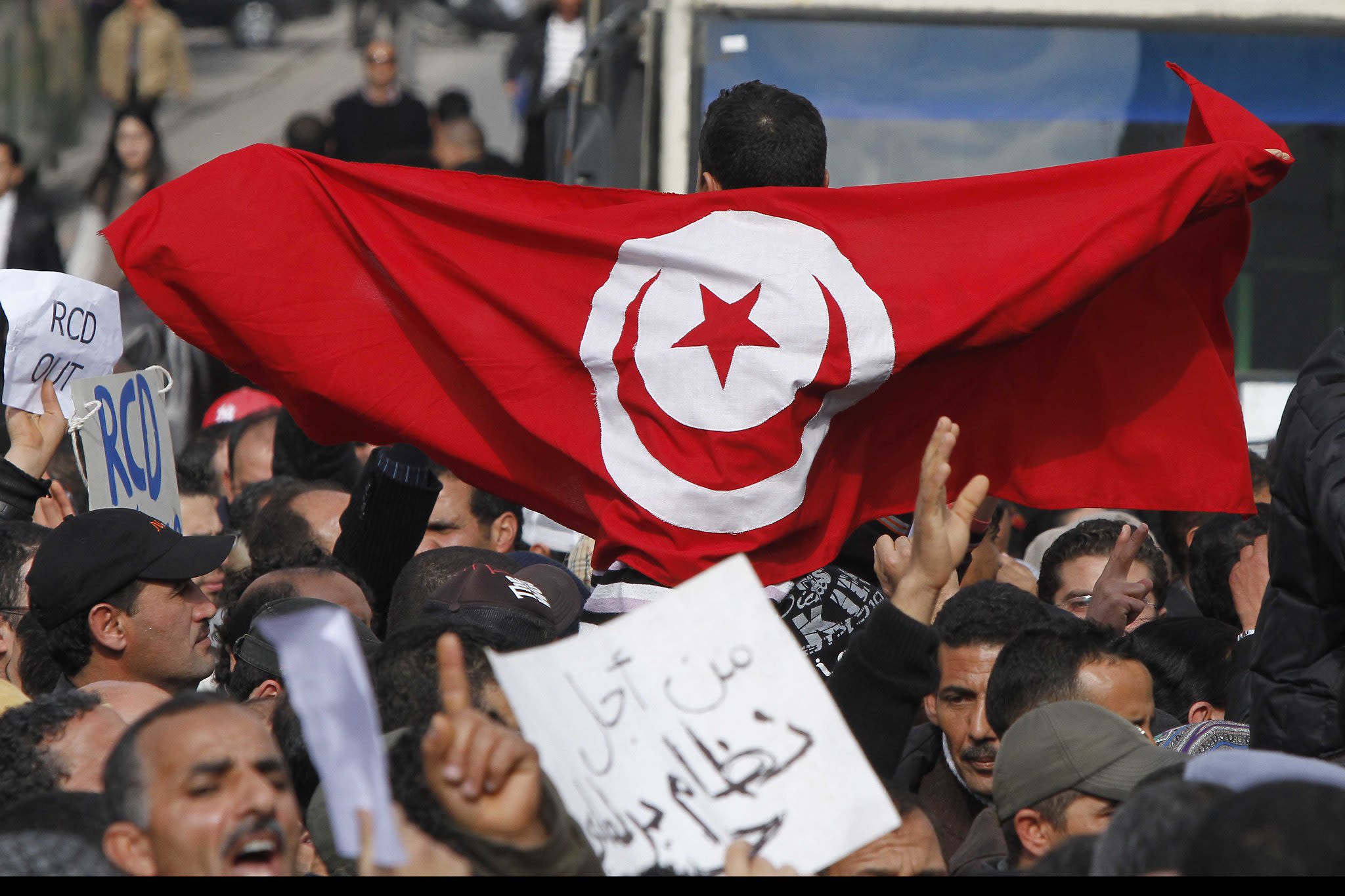 A crowd of people at a street protest. Some are holding signs. One has the Tunisian flag around his shoulders.