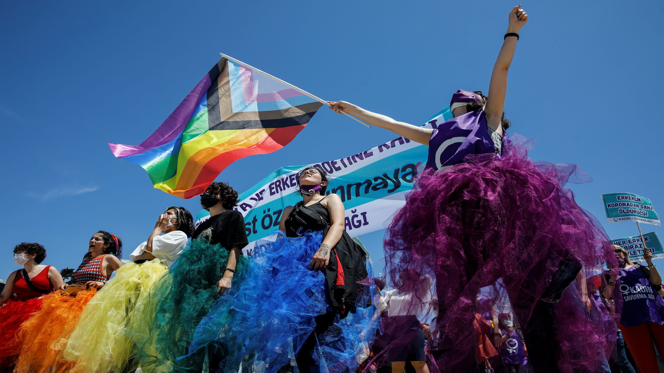A group of people wearing rainbow-coloured skirts and flying the LGBT pride flag against a bright blue sky.